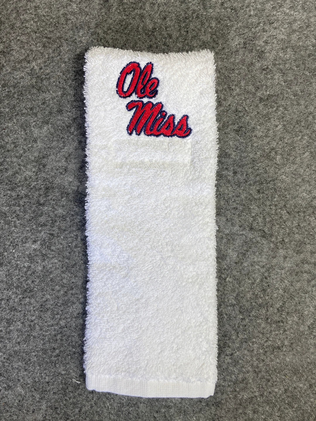 A white terry cloth towel with 'Ole Miss' embroidered in red and blue on the top, laid flat on a gray textured background.